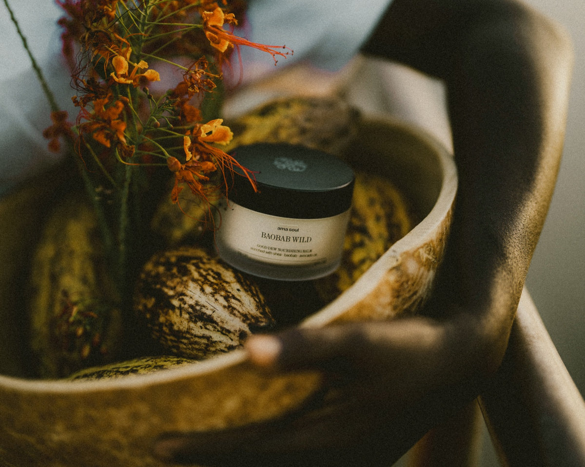 Ama Soul Baobab Wild Good Dew balm in a carved wooden bowl with cocoa pods and orange botanicals